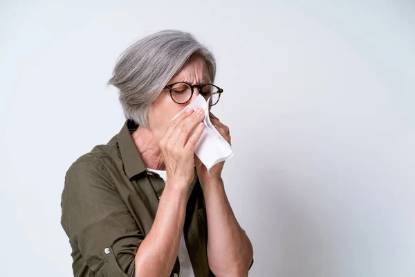 Nose running or flue, mature woman blow her nose in napkin. Senior grey haired woman suffering from covid, coronavirus. Online doctors for mature people. Flue sick senior woman.