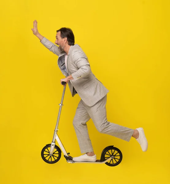 Waving hand handsome middle aged man in white casual suit ride scooter looking sideways. Mobility for middle aged business man isolated on yellow background. Business concept.