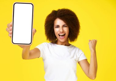 En iyi teklif, elini kaldırdığı için mutlu olan heyecanlı Afro-Amerikalı kadın sarı arka planda beyaz tişört giyen büyük, beyaz ekranlı akıllı telefonu tut. Uygulama reklamı.. 