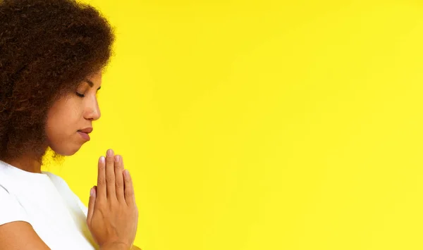 Young african american woman in prayer position wearing white t-shirt isolated on yellow background. African girl praying God studio shot. Spiritual woman put hands in prayer.