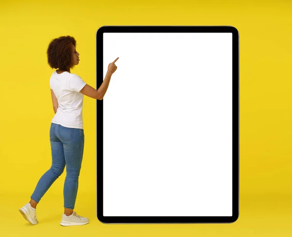 African american young woman with afro hair stand in front big smartphone with white screen use touchscreen wearing white t-shirt isolated on yellow background. Great offer. App advertisement.