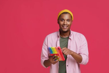 Yakışıklı Afro-Amerikan genç adam, gökkuşağında dijital tablet kullanan ya da sosyal medyada mutlu bir şekilde gülümseyen pembe tişört ve sarı şapka kullanan adam. Çevrimiçi randevu kavramı. 