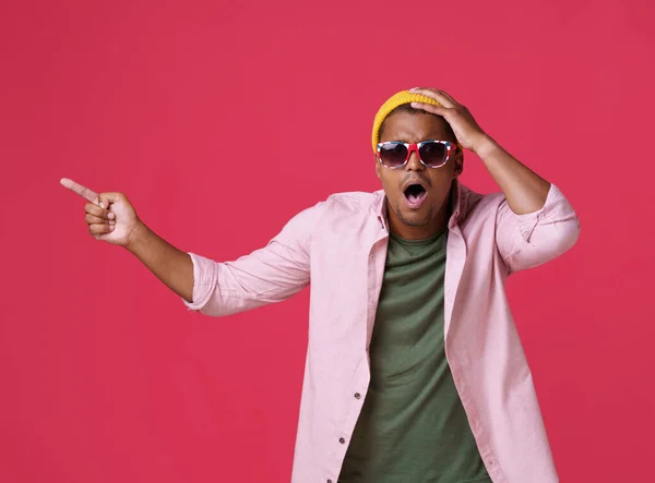 Pointing sideways stretched young african american man in UK, Britain flag sunglasses, wearing casual excited or shocked touching his head in yellow hat isolated on red background.