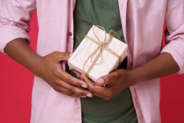 Close up on man hands with gift. African American guy with present in hands wrapped in crafted paper wearing casual pink shirt and green t-shirt. no face visible.