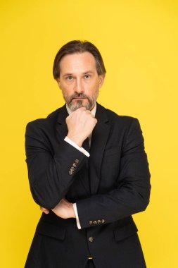 Siyah takım elbiseli, zarif, yakışıklı, orta yaşlı bir adam. Elleri katlı ve sarı arka planda izole edilmiş kameraya bakıyor. Siyah takım elbiseli yetkili iş adamı.. 