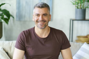 Rahat, orta yaşlı, rahat, gri saçlı, rahat bir adam kanepede oturuyor. Evde mutlu ve olgun bir adam var. Kanepede oturan serbest çalışan adam.. 