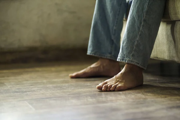 Close up on a male bare feet on the hard wood floor while sitting on the sofa in semi light room wearing denim jeans. Man bare feet on the floor. No face visible.