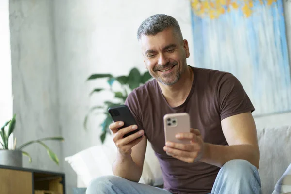 Using two modern smartphones sitting on the couch handsome grey haired middle aged freelancer man working at home. Setting up mobile phone man sits at home. Business from home concept.