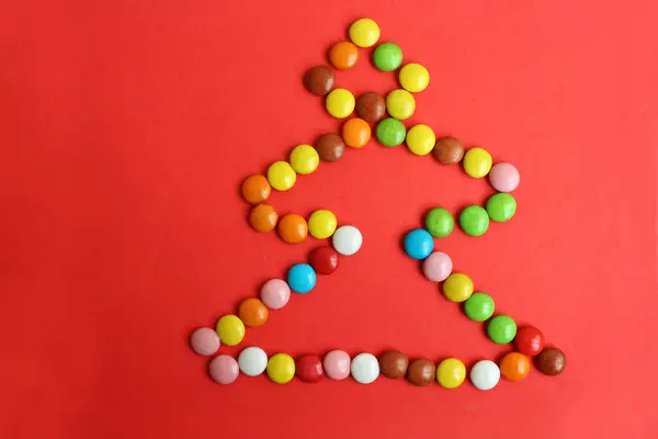 Petits bonbons au chocolat sont disposés sous la forme d'un arbre de Noël sur un fond rouge. Un symbole de la nouvelle année et de Noël. Vue de dessus. Les bonbons sont multicolores. Espace de copie pour le texte. — Photo