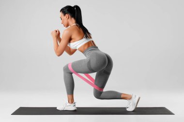 Atletik kız gri arka planda direnç bandıyla kalça egzersizi yapıyor. Spor salonundaki kadın.