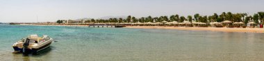 Hurghada, Mısır - 22 Eylül 2021: Motorlu tekneden ve Kızıl Deniz kıyısından panoramik manzara. İnsanlar yeşil palmiye ağaçlarıyla kumlu bir sahilde dinleniyor ve yüzüyorlar..