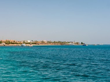 Hurghada, Mısır - 22 Eylül 2021: Kızıl Deniz 'in gök mavisi suyu ve yeşil palmiye ağaçlı kumlu Mısır plajı. Tekneler ve yatlar kıyıya yakın duruyor. Boşluğu kopyala.