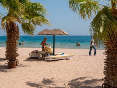 Hurghada, Mısır - 22 Eylül 2021: Kadın palmiye ağaçlarının yanındaki saman şemsiyesinin altına bir güneş şezlonguna havlu seriyor. Fiyat listesi olan bir adam sahilde gezintiler teklif ediyor. İnsanlar Kızıl Deniz 'de yüzer..