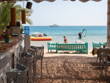Hurghada, Mısır - 22 Eylül 2021: Kızıldeniz sahilindeki barın görüntüsü yüzen insanların arka planına karşı. Uzun, gri, tahta sandalyeler barın yanında..