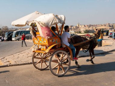 Giza, Kahire, Mısır - 30 Eylül 2021: Mısır piramitlerinin en büyüğü olan Cheops Piramidi. Mısırlılar Kahire şehrinin arka planına karşı at arabasıyla yol boyunca ilerliyorlar..
