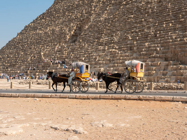 Giza, Cairo, Egypt - September 30, 2021: Pyramid of Cheops, the largest of the Egyptian pyramids. Tourists walk, take pictures and ride a cart with a horse along the road along the pyramid.