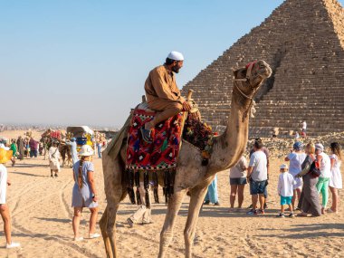 Giza, Kahire, Mısır - 30 Eylül 2021: Mısırlı milli elbiseli bir deve üzerinde oturuyor. Turistler Mısır piramidinin yakınında yürüyüp fotoğraf çekiyorlar.
