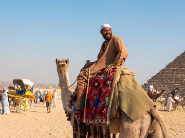 Giza, Kahire, Mısır - 30 Eylül 2021: Mısırlı milli elbiseli bir deve üzerinde oturuyor ve gülümsüyor. Turistler Mısır piramidinin yakınında yürüyüp fotoğraf çekiyorlar.
