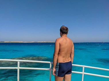 Hurghada, Mısır - 1 Ekim 2021: Yüzme şortlu bir adam yatta duruyor ve gök mavisi kızıl denize bakıyor. Boşluğu kopyala.