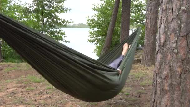 Een man of vrouw ligt in een hangmat aan de oever van een bosmeer. Wandelen ontspannen — Stockvideo