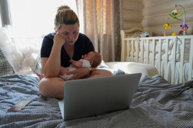 Kollarında çocuğu olan genç bir kadın, kafası karışmış bir dizüstü bilgisayarın yanında oturuyor.