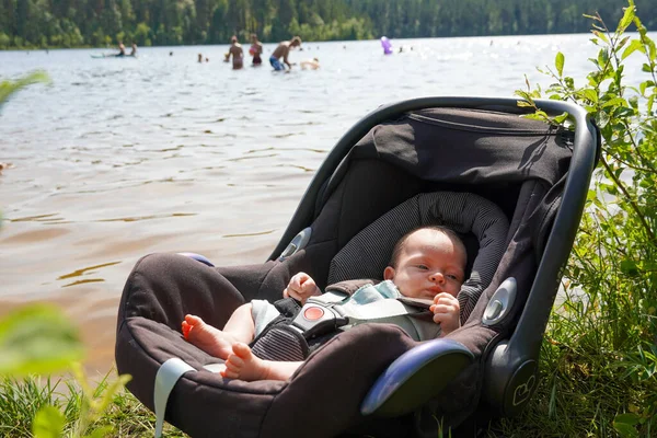 Newborn baby in a child car seat on the shore of the lake with people bathing 图库图片