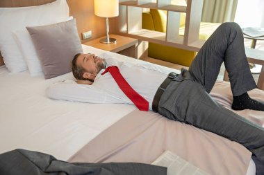 Takım elbiseli bir işadamı otel yatağında uzanıyor, dinleniyor.