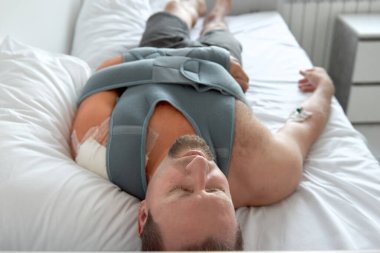 Hastane koğuşunda yatağında kateter takmış bir adam var. Elastik omuz kırığı. Acil durum.