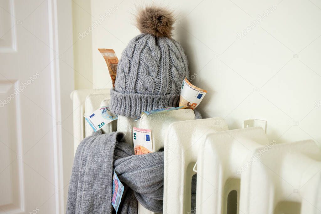 Vintage heating radiator with winter hat and scarf stuffed with euro money. The electricity bill goes up, European energy crisis concept. 