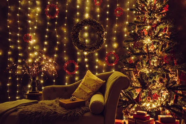 Festive decorated home interior living room in with lot of string LED lights, led curtains and pine cone and berry wreaths on wall, red Christmas gifts under the tree, comfortable canape sofa waiting.