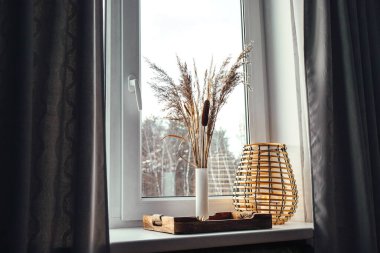 Using dried natural various grass plants inside vase indoors for home decoration. Vase with various plants and wood black metal wire lantern on window sill in daylight.  clipart