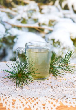 Masadaki şeffaf cam çay fincanında sıcak çam ağacı iğnesi çayı. Arka planda karlı çam ağacı, soğuk kış gününde açık havada.