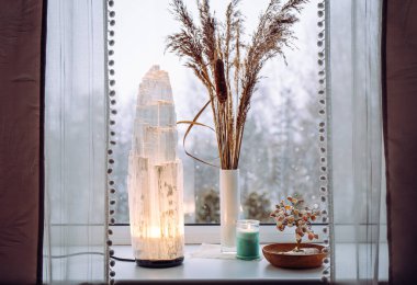 Rough big selenite crystal tower pole lamp illuminated on home window sill, spiritual home decor accent. Winter forest on background. clipart