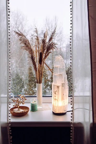 Rough big selenite crystal tower pole lamp illuminated on home window sill, spiritual home decor accent. Winter forest on background.