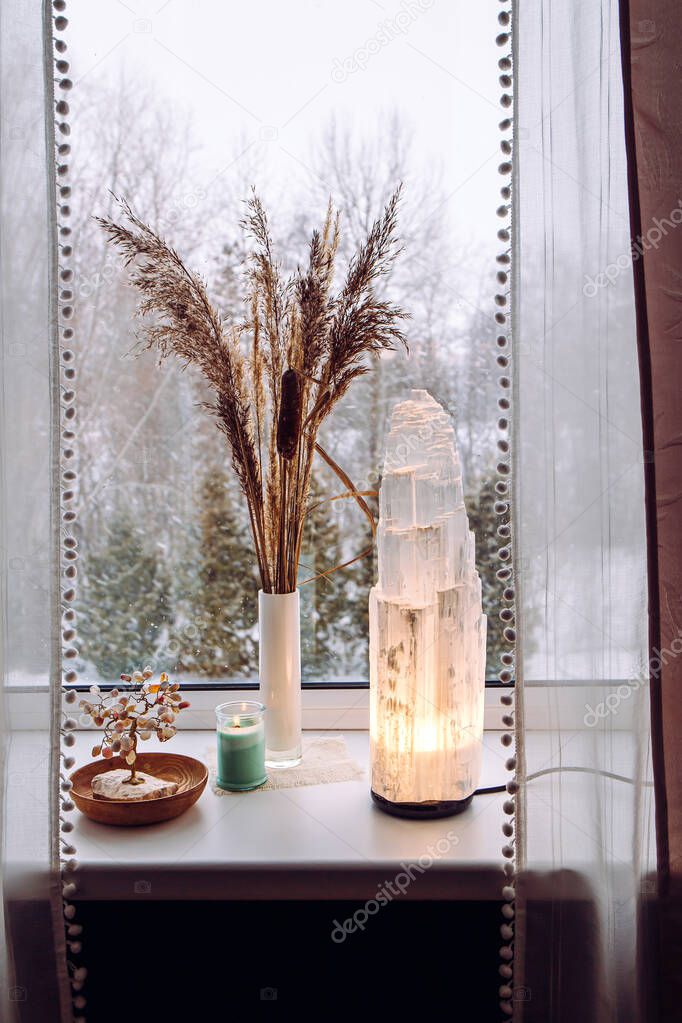 Rough big selenite crystal tower pole lamp illuminated on home window sill, spiritual home decor accent. Winter forest on background.