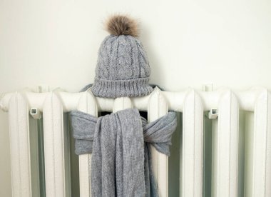 Gri yün örülmüş, kışlık şapka ve eşarplı klasik ısıtma radyatörü. Elektrik ve doğalgaz faturası yükseliyor, Avrupa enerji krizi konsepti. Boşluğu kopyala, metin için yer aç.