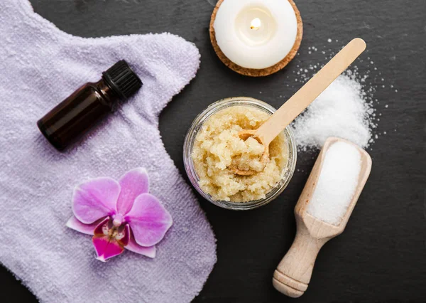 Homemade sugar body scrub in glass jar, decorated with orchid blossom, body scrub glove wooden spoon with sugar powder on black stone cutting board. Body skin care concept.