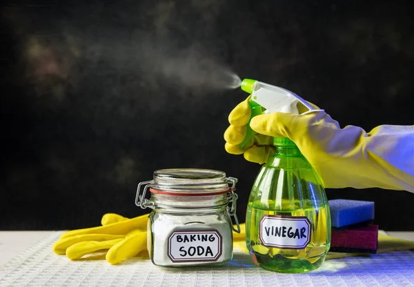 Using baking soda Sodium bicarbonate and white vinegar for home cleaning. White vinegar in spray bottle and baking soda in glass jar. Person spraying mist from bottle.