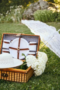 Romantic set with vintage picnic basket, lace sun umbrella and summer hat on park garden lawn on sunny summer day outdoors.