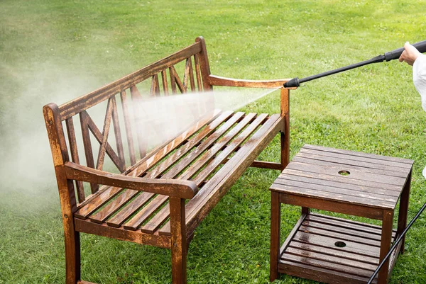 Close up view of person working cleaning pressure washing wooden garden furniture bench outdoors in summer.