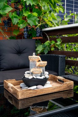 Home patio with black plastic garden furniture, small relaxing electrical zen table fountain on table and real grape vines with grapes hanging on background. Home decor.