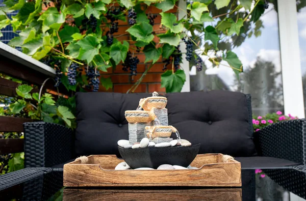 Home patio with black plastic garden furniture, small relaxing electrical zen table fountain on table and real grape vines with grapes hanging on background. Home decor.