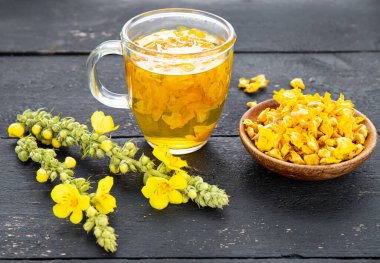 Herbal medicinal tea drink made of Verbascum thapsus, the great mullein, greater mullein or common mullein. Yellow dried flower petals. Glass cup with tea on black background and dried petals in bowl.