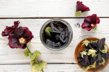 Herbal medicinal tea drink made of Malva sylvestris L. Known as common mallow, cheeses, high mallow and tall mallow. Glass cup with tea on white background fresh and dried petals scattered.