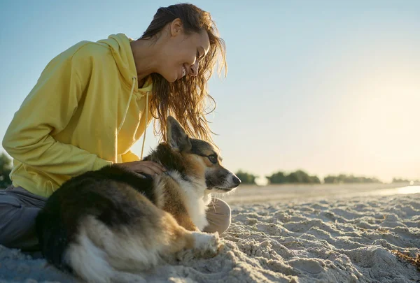 ผู้หญิงที่มีความสุขยิ้มกับสัตว์เลี้ยงของเธอ Welsh Corgi สุนัขใช้เวลาร่วมกันที่ชายหาดแดด — ภาพถ่ายสต็อก