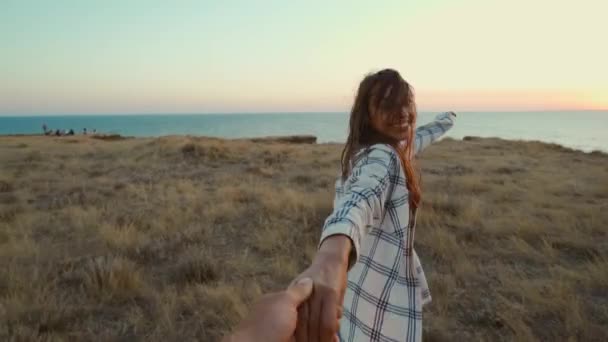 Счастливые положительные эмоции девушка смеется и держит за руку своего друга на скале с морской накидкой во время заката. — стоковое видео