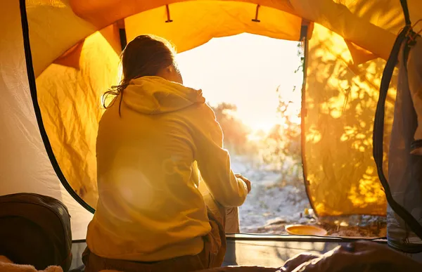 Matkailija nainen teltassa ihaillen aamuauringonnousua, näkymä takaapäin — kuvapankkivalokuva