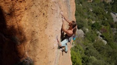 Güçlü yakışıklı kaya tırmanışçısının yavaş çekim üst görüntüsü zorlu yoldan dikey uçuruma tırmanır. Atletik tırmanışçı elle sıkı çaba sarf eder, kayaya uzanır ve tutunur, dinlenir ve yukarı bakar.