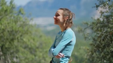 Doğal yaz arka planında neşeli, genç ve sportif kadın kaya tırmanışçısının yan görünüm portresi. Kız gülümsüyor, gülüyor, arkadaşlarıyla konuşuyor ve bazen yukarı bakıyor.