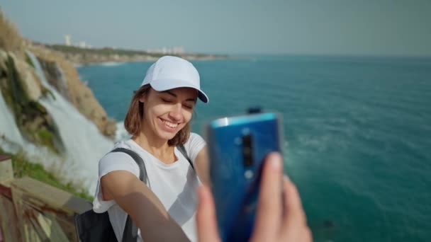 Atractiva mujer sonriente turista haciendo selfie en el teléfono móvil en el fondo de la cascada Duden inferior en Antalya. Famoso punto de vista en la costa del mar Mediterráneo en Turquía. — Vídeos de Stock